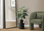Artificial Medium Alocasia plant in a black pot on a pedestal next to a green armchair and floor mirror.