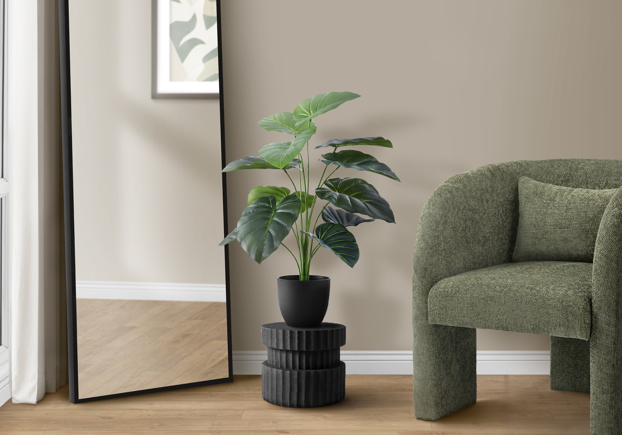 Artificial Medium Alocasia plant in a black pot on a pedestal next to a green armchair and floor mirror.