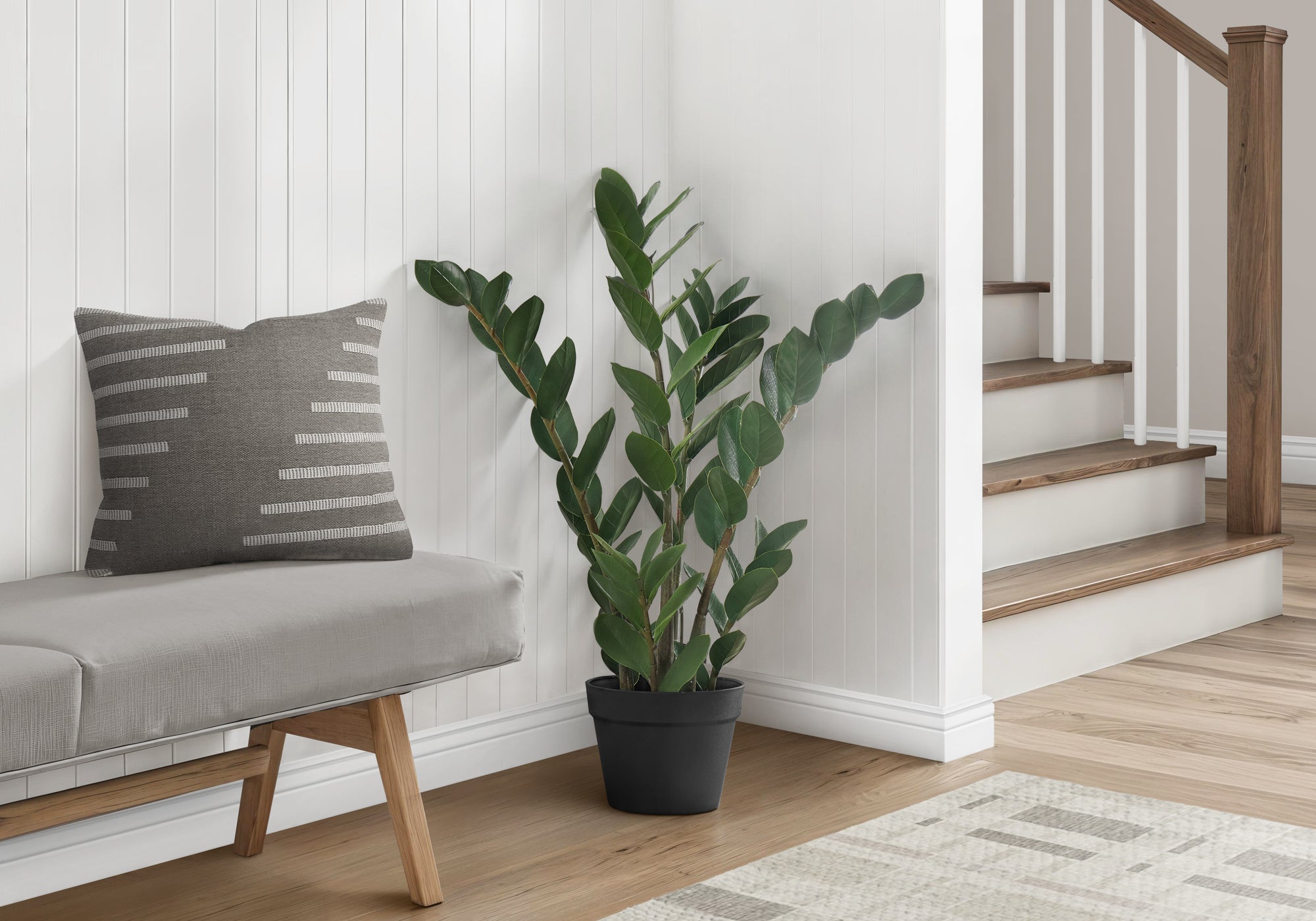 A medium artificial ZZ plant in a black pot sits on a wooden floor next to a grey bench with a patterned pillow.