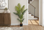 A tall artificial Majesty Palm tree in a black pot stands on a wooden floor next to a wooden cabinet in a bright home.