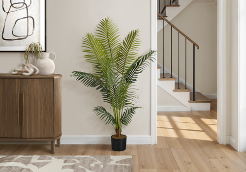 A tall artificial Majesty Palm tree in a black pot stands on a wooden floor next to a wooden cabinet in a bright home.