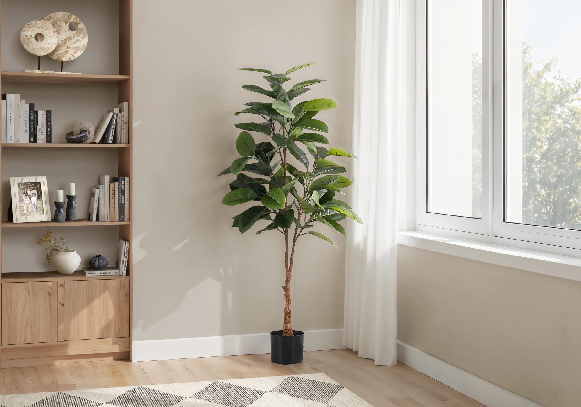 A tall artificial rubber tree in a black pot stands in a bright room next to a wooden bookshelf and a window.