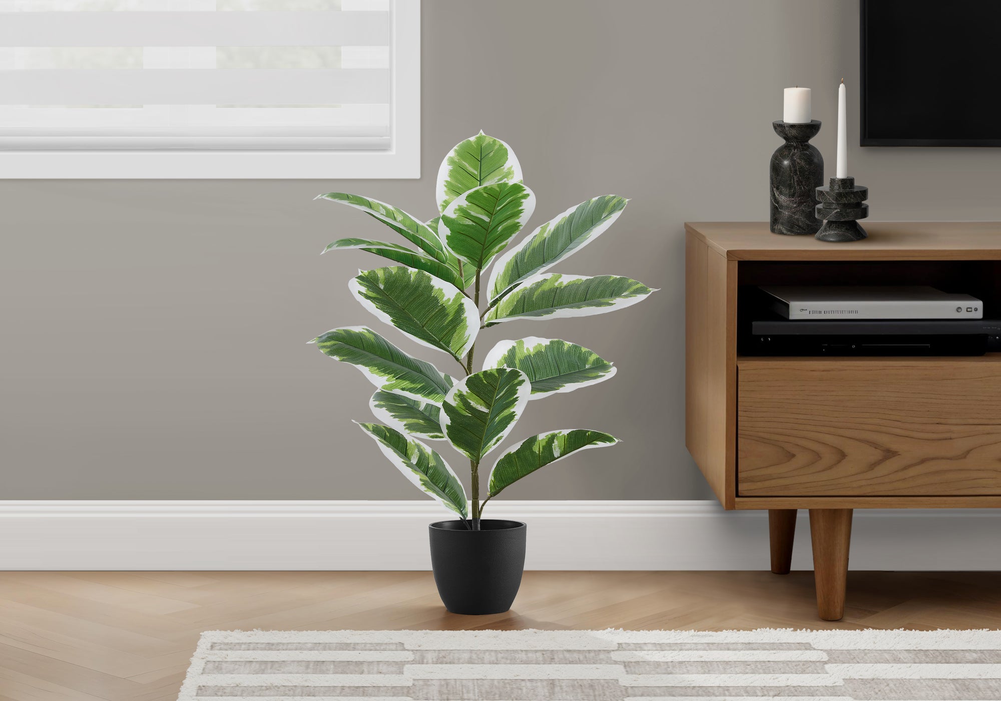 A medium artificial variegated rubber tree in a black pot sits on a wooden floor beside a wooden media console.
