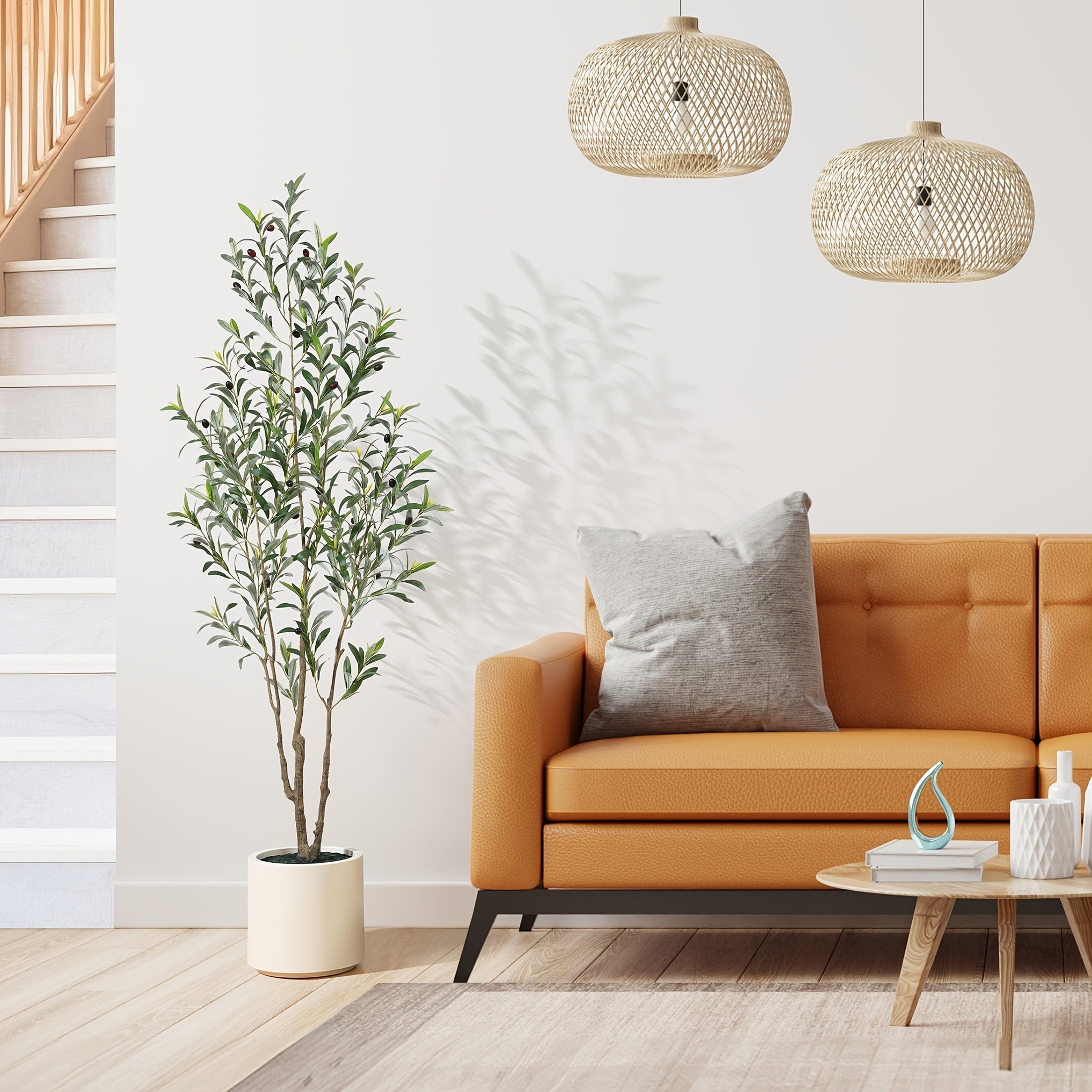 A tall artificial olive tree in a white pot stands next to a tan leather sofa in a bright living room.
