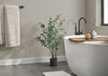 A medium artificial eucalyptus plant in a black pot stands next to a white freestanding bathtub in a modern bathroom.
