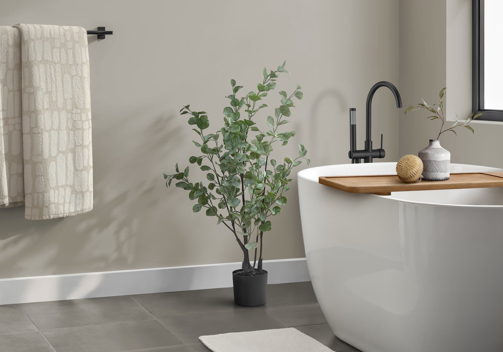 A medium artificial eucalyptus plant in a black pot stands next to a white freestanding bathtub in a modern bathroom.