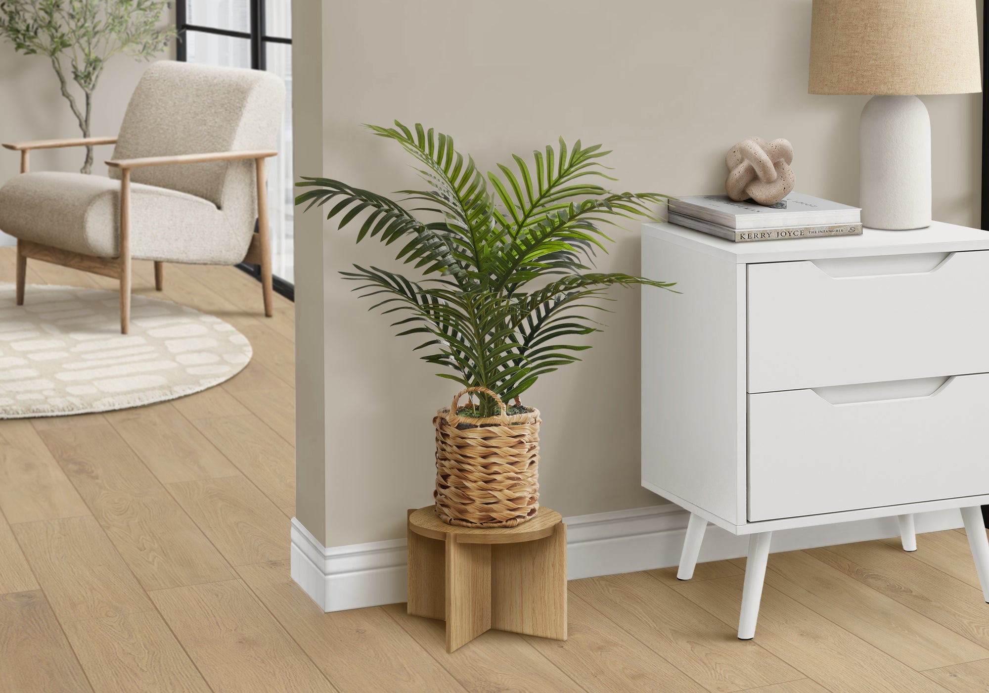 An artificial parlor palm in a woven basket sits on a wooden stand next to a white dresser in a neutral room.