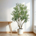 A large artificial olive tree with green leaves and small black olives in a white pot standing next to a wooden table.