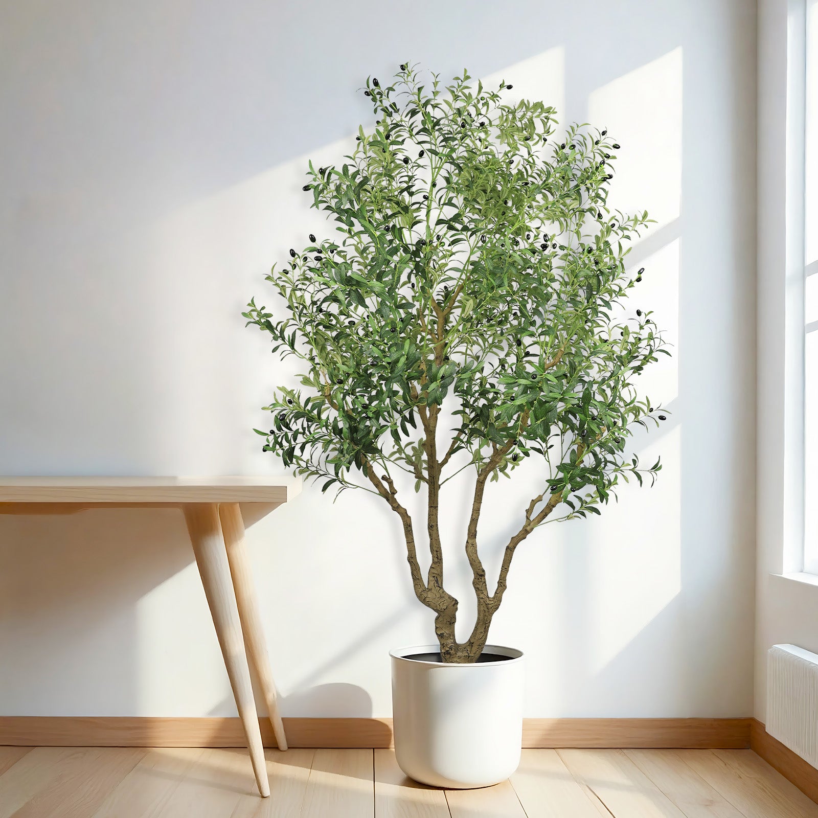 A large artificial olive tree with green leaves and small black olives in a white pot standing next to a wooden table.