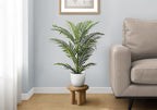 A medium artificial Areca palm in a white pot, sitting on a small wooden stool next to a beige sofa in a living room.