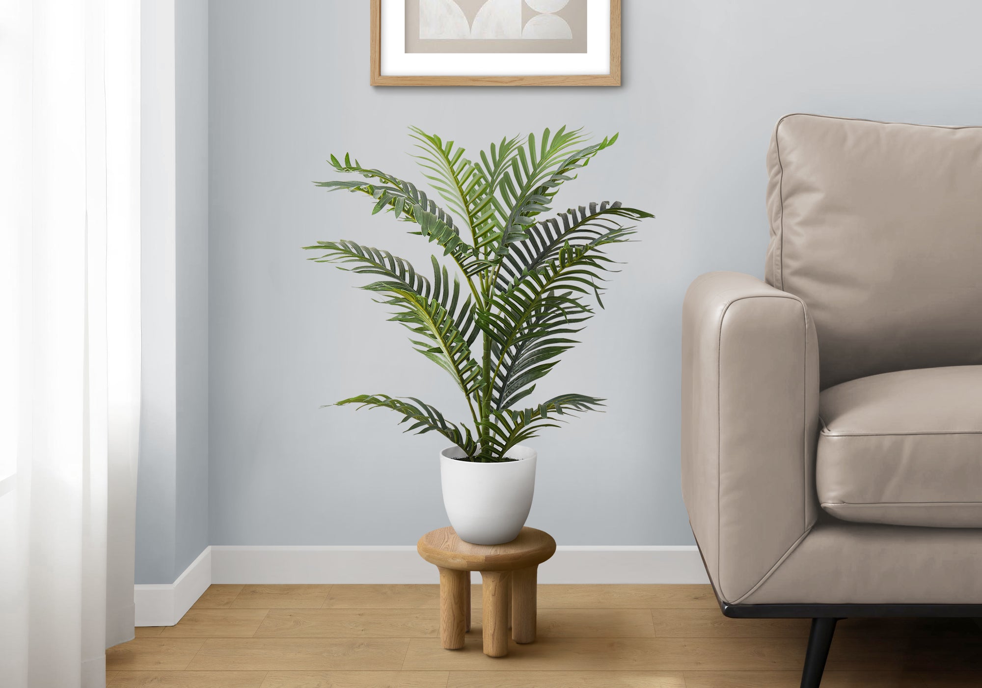 A medium artificial Areca palm in a white pot, sitting on a small wooden stool next to a beige sofa in a living room.