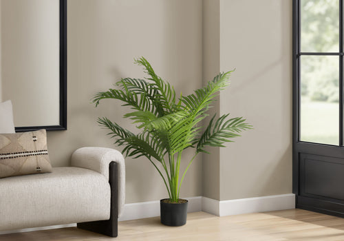An artificial Cat Palm with wide, green fronds in a black pot, placed in a corner next to a modern armchair.