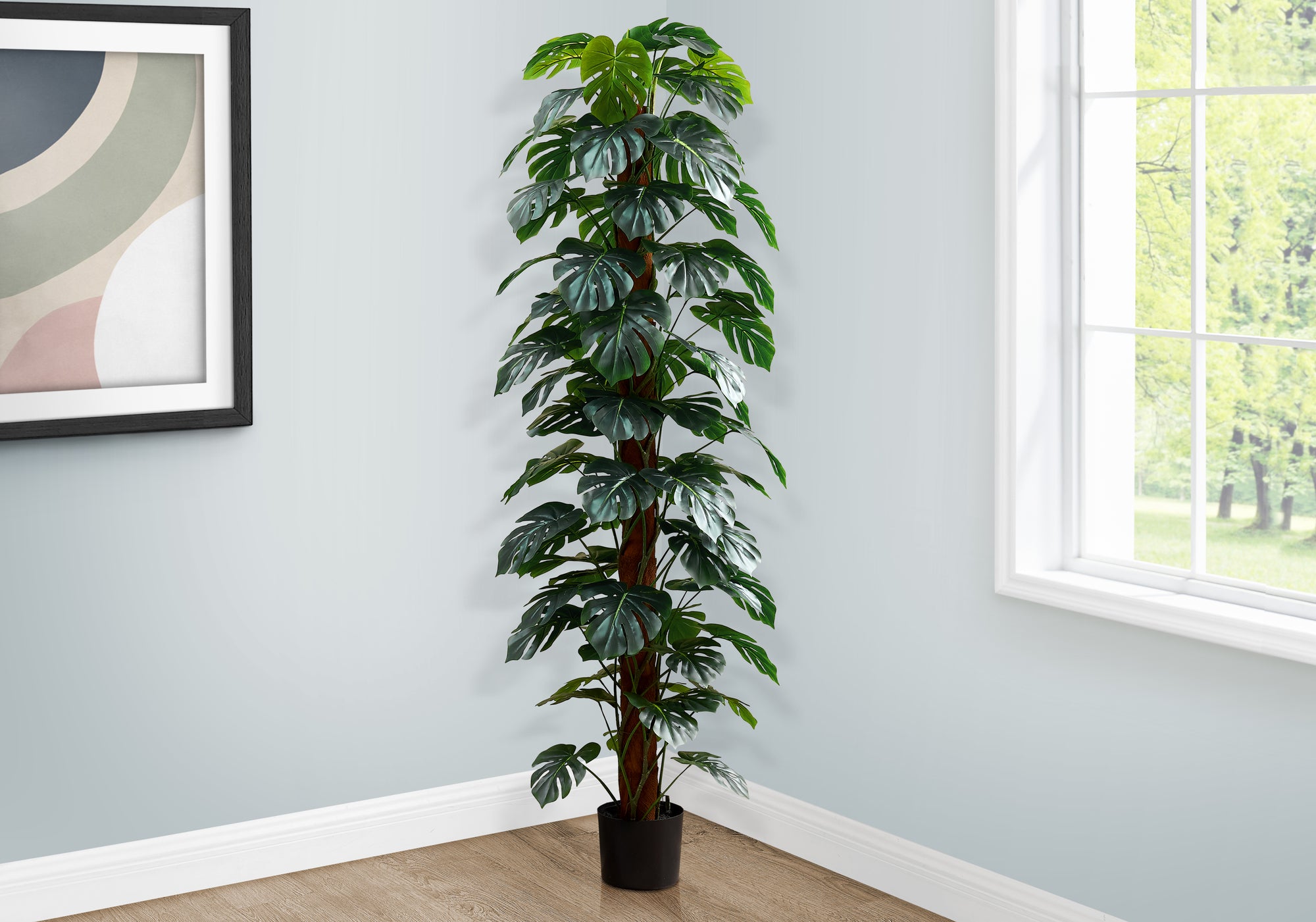 Tall artificial Monstera plant with split leaves climbing a moss pole, placed in the corner of a bright room.