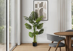 An artificial Monstera plant in a black pot stands in a modern room corner next to a glass sliding door.
