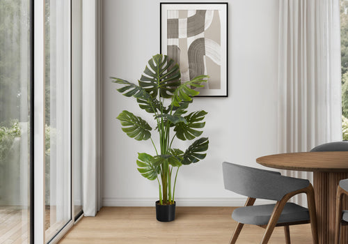 An artificial Monstera plant in a black pot stands in a modern room corner next to a glass sliding door.