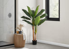A tall artificial banana plant with large green leaves stands in a corner next to a fluted wooden side table.