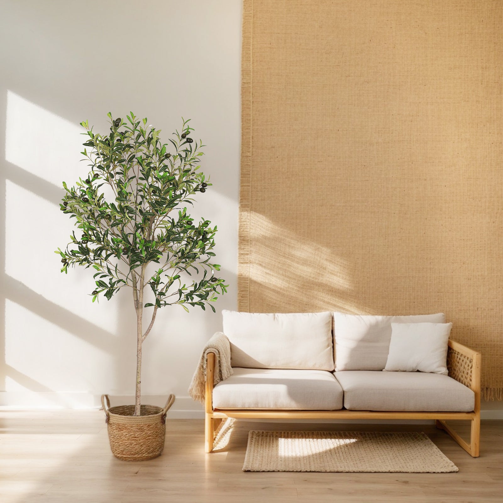 A tall artificial olive tree in a woven basket stands next to a light-colored wooden sofa in a sunlit room.