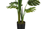 Close-up of an artificial Monstera plant showing thick green stems and split leaves in a black pot.