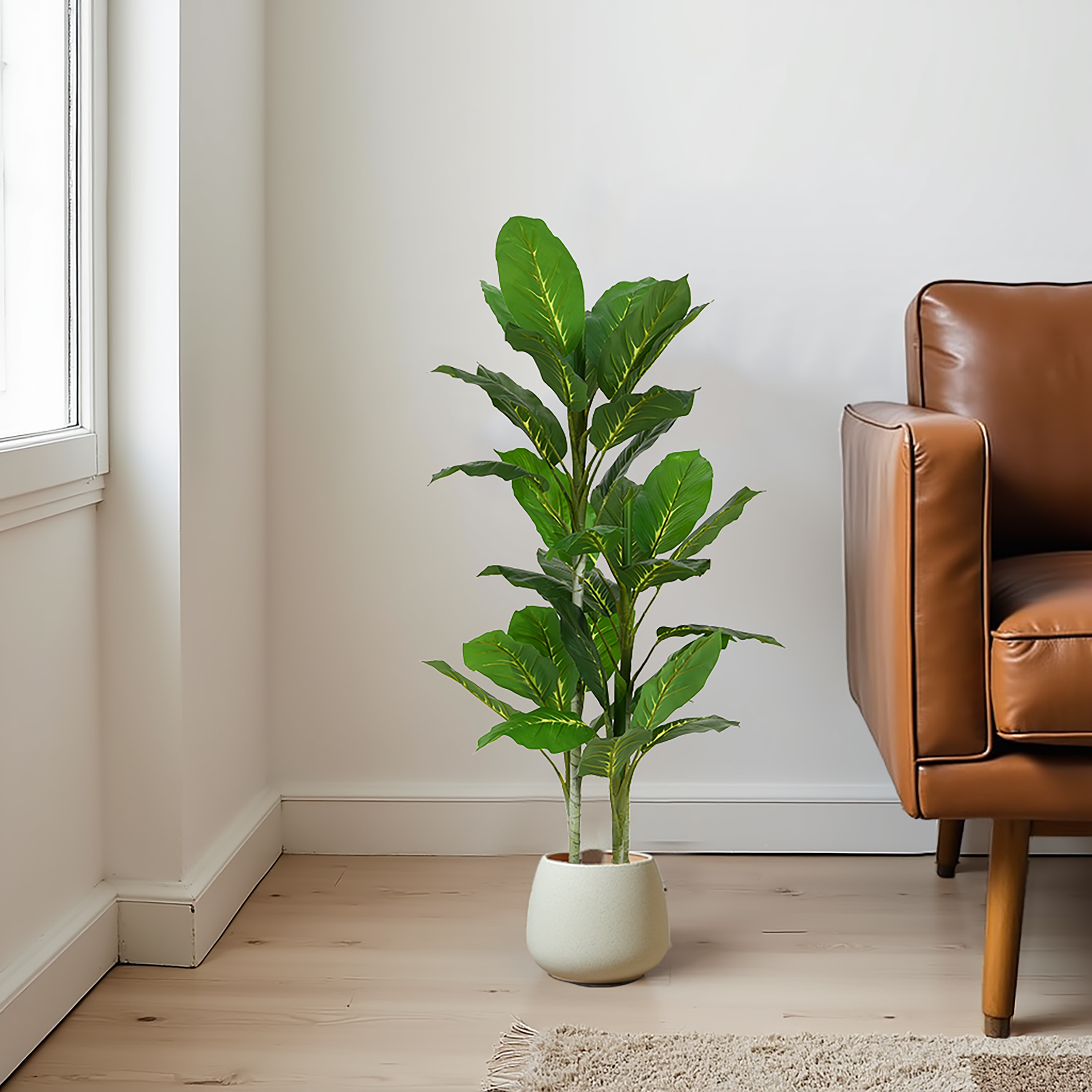 A tall artificial Dieffenbachia plant with broad, patterned green leaves in a light-colored pot, placed next to a brown leather sofa.