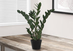 A medium artificial ZZ plant with dark green leaves in a black pot, sitting on a rustic wooden desk near a window.