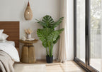 Large artificial Monstera plant in a black pot standing next to a brass side table in a bright bedroom.
