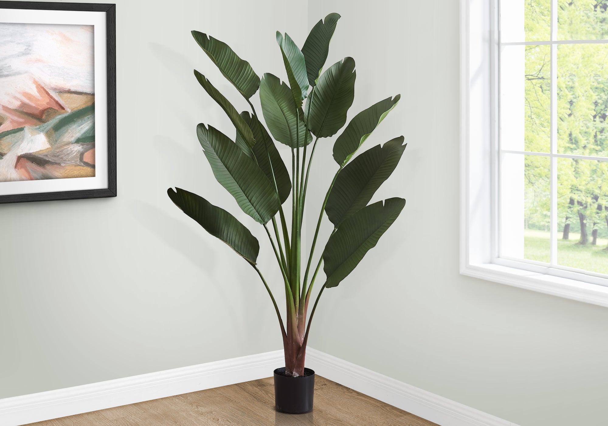 A tall artificial Bird of Paradise plant with large green leaves in a black pot, placed in a bright room corner.