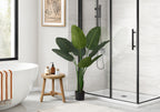 A large artificial Bird of Paradise plant in a black pot stands in a modern bathroom next to a wooden stool.