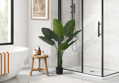 A large artificial Bird of Paradise plant in a black pot stands in a modern bathroom next to a wooden stool.