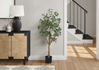 A tall artificial eucalyptus tree in a black pot standing on a wooden floor next to a textured cabinet and staircase.