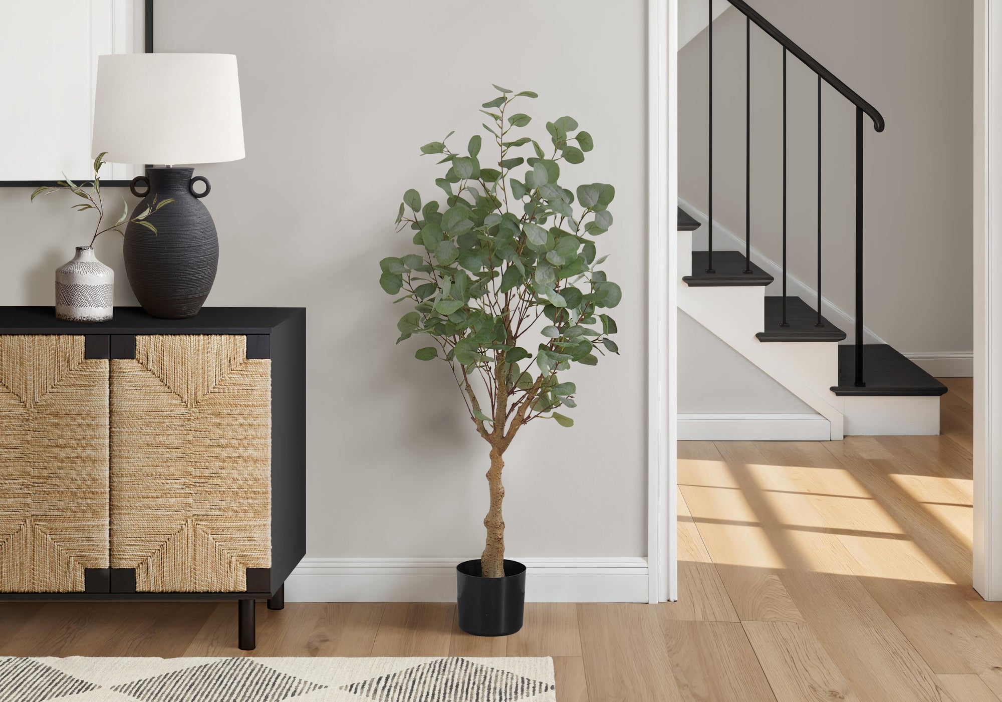 A tall artificial eucalyptus tree in a black pot standing on a wooden floor next to a textured cabinet and staircase.