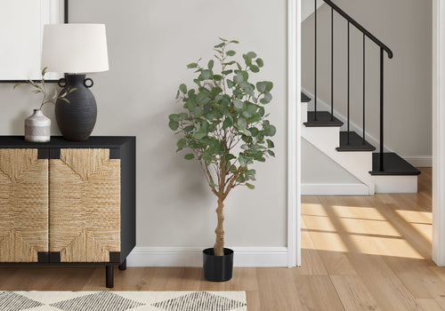 A tall artificial eucalyptus tree in a black pot standing on a wooden floor next to a textured cabinet and staircase.