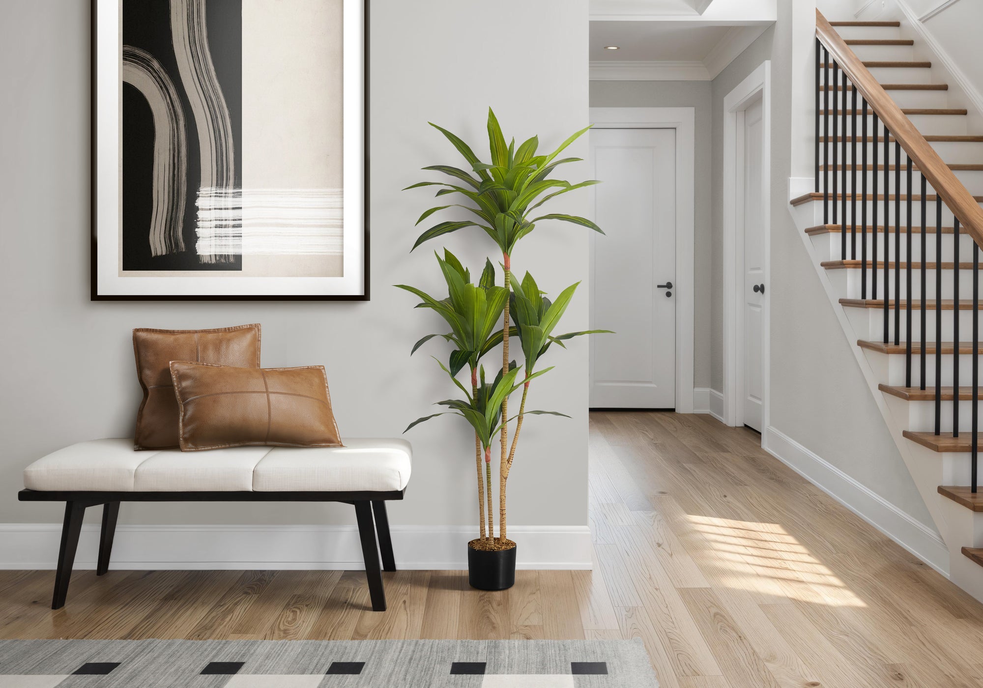 A tall faux Dracaena tree with three trunks stands in a bright entryway next to a white bench with leather pillows.