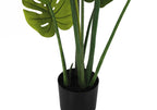 Close-up of an artificial Monstera plant with large, split green leaves in a simple black pot against white.