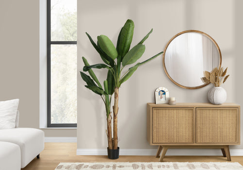 A tall artificial banana plant with large green leaves stands in a living room next to a wooden cane sideboard.