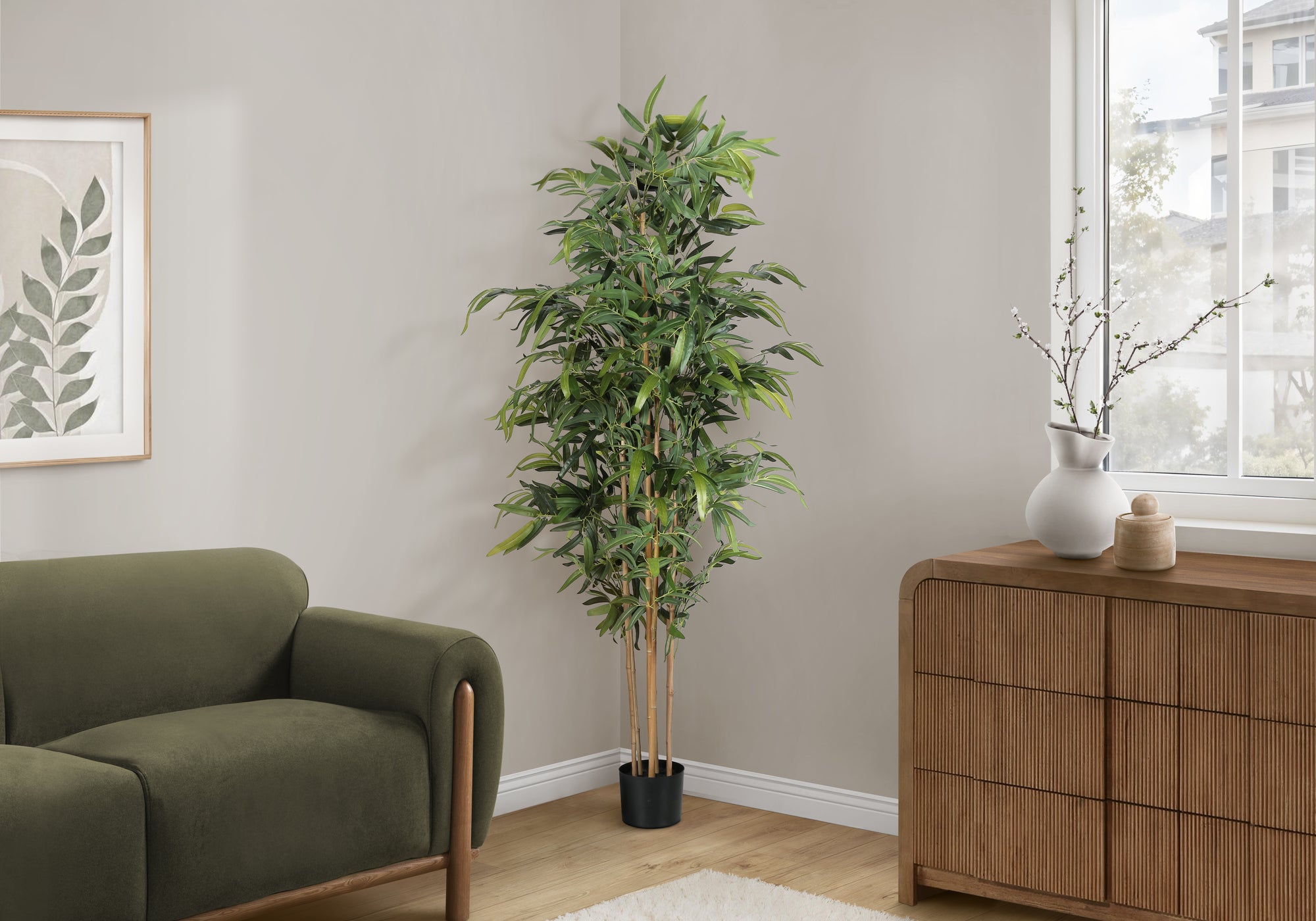 A tall artificial bamboo tree in a black pot standing in the corner of a living room next to a green sofa.