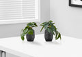 Two small artificial plants in black and white patterned pots sitting on a white table near a window.