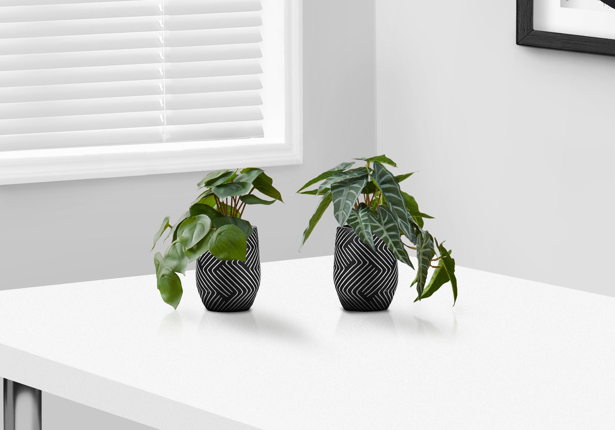 Two small artificial plants in black and white patterned pots sitting on a white table near a window.