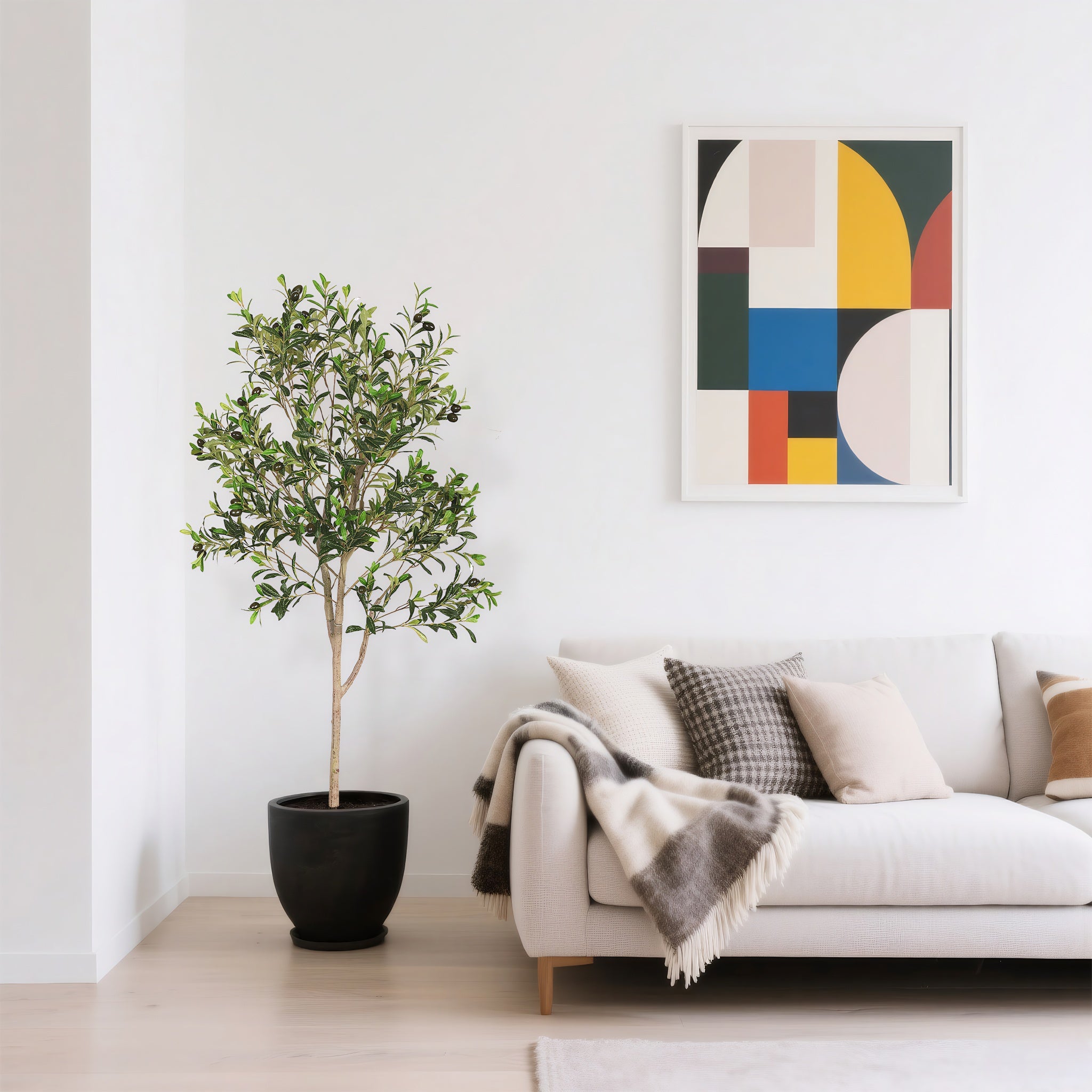 A five-foot artificial olive tree in a black pot stands next to a white sofa with patterned pillows and a throw blanket in a bright living room.