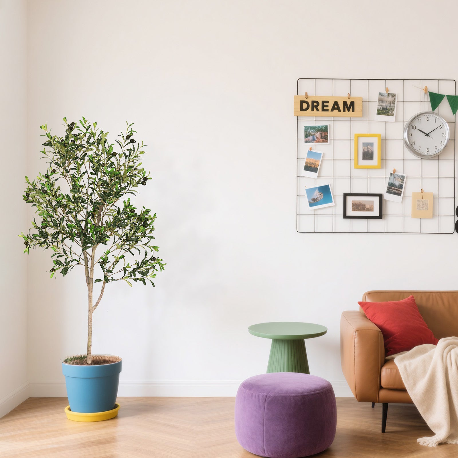 A five-foot artificial olive tree in a blue pot standing in a living room next to a brown leather sofa and purple ottoman.