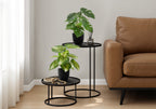 Two small artificial potted plants on black nesting tables next to a brown leather sofa in a living room.