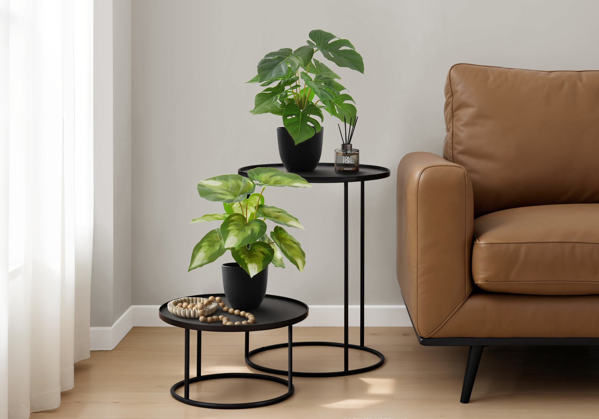 Two small artificial potted plants on black nesting tables next to a brown leather sofa in a living room.