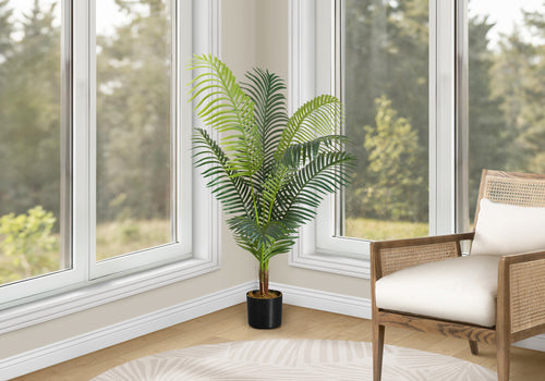 A large artificial Majesty Palm tree in a black pot stands in a corner between two windows next to a wooden chair.