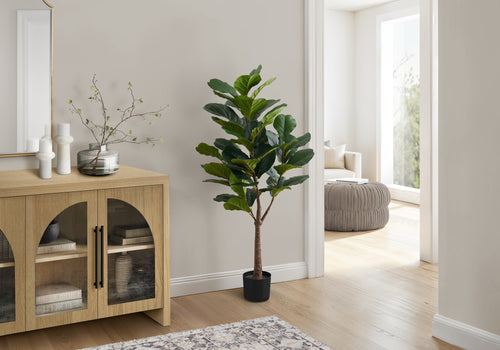 A large artificial fiddle leaf fig tree in a black pot standing next to a light wood cabinet in a bright living room.