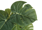 Close-up of the textured green leaves of an artificial Monstera plant against a white background.
