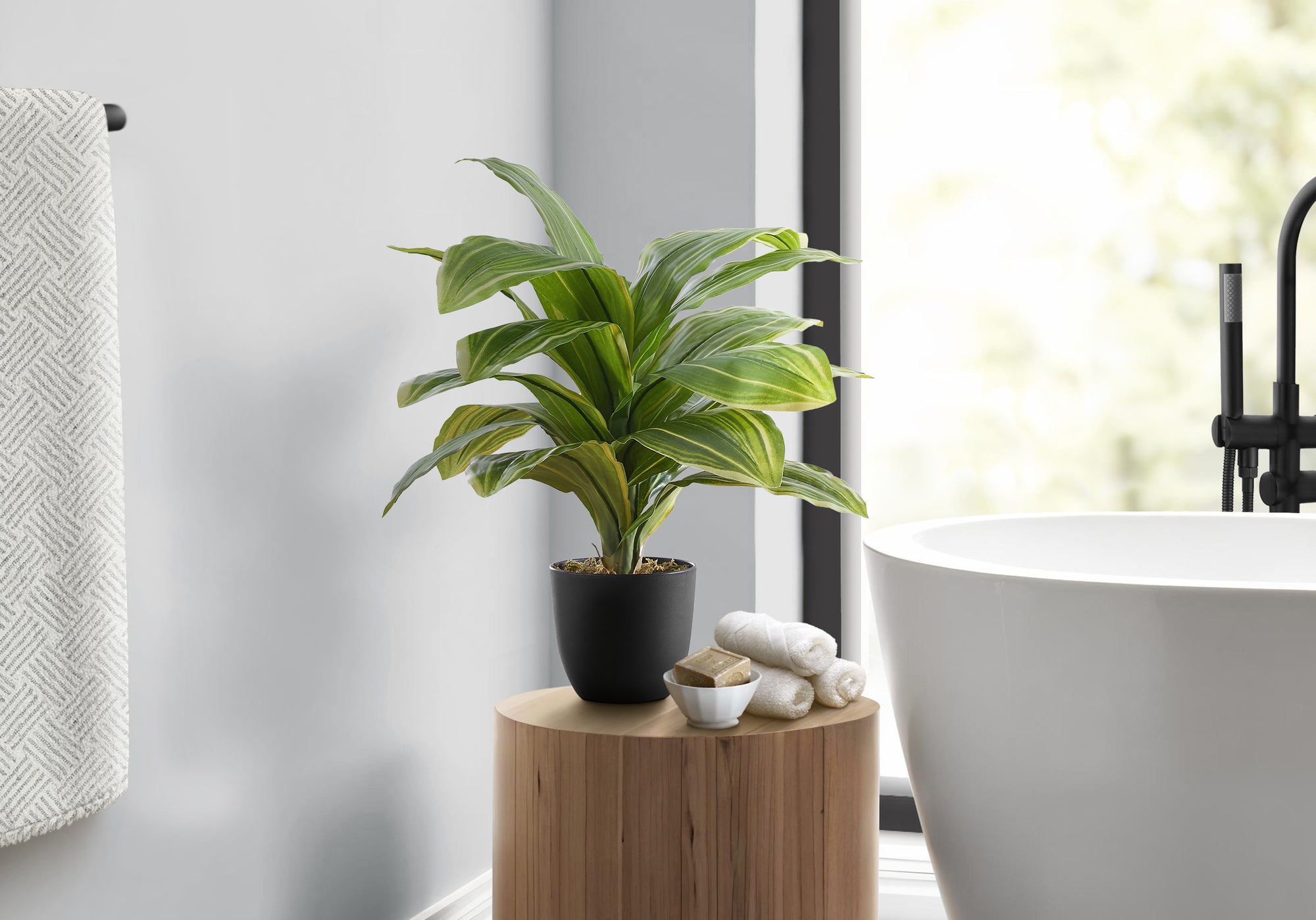 A small faux Dracaena Lemon Lime plant in a black pot sits on a wooden stool next to a white bathtub in a bathroom.