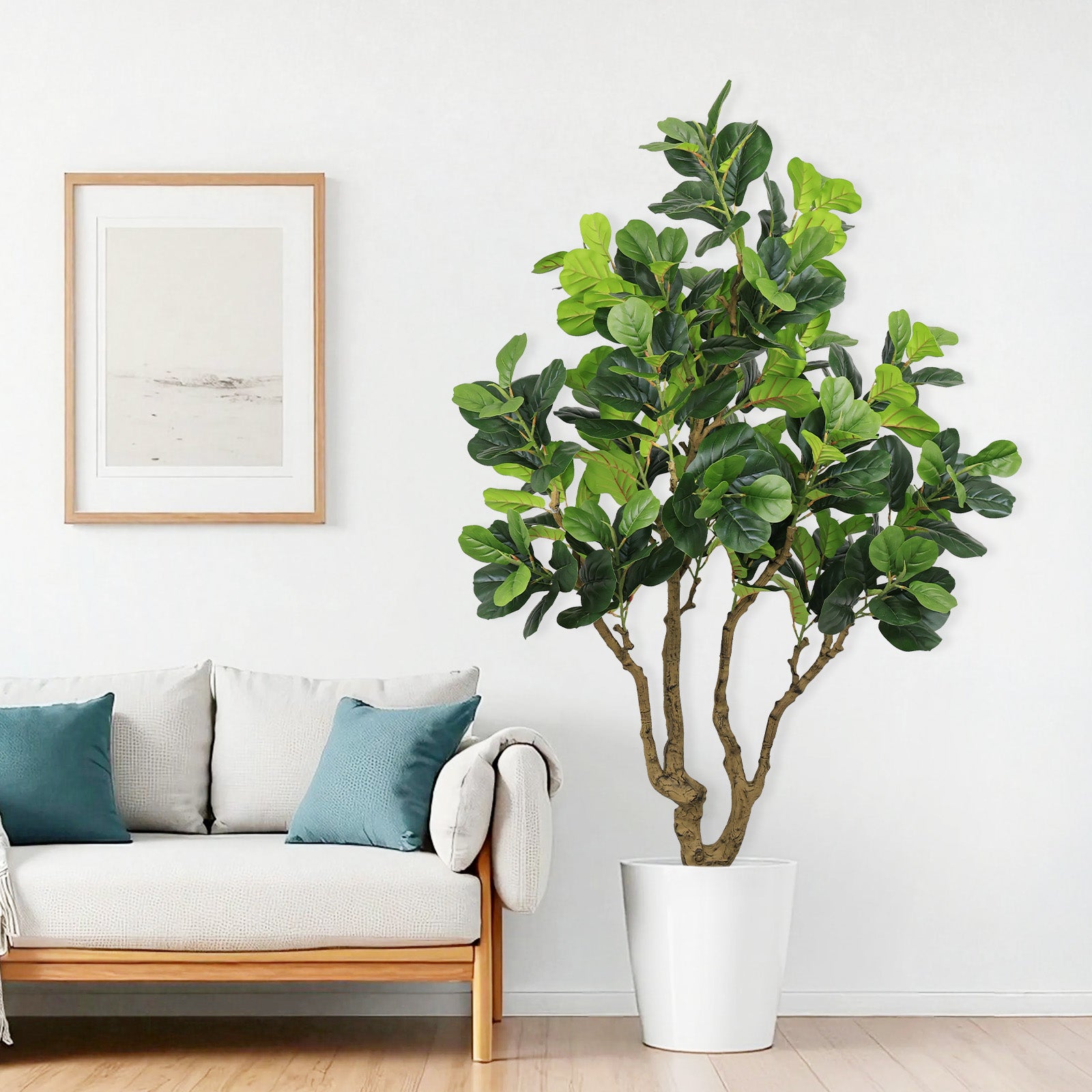 A tall artificial Fiddle Leaf Fig tree in a white pot standing next to a light-colored sofa with teal pillows.