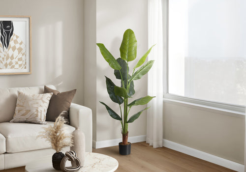 A tall artificial Bird of Paradise plant in a black pot standing in a bright living room corner next to a sofa.