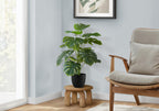An artificial Medium Monstera plant in a black pot on a small wooden stool next to an armchair.