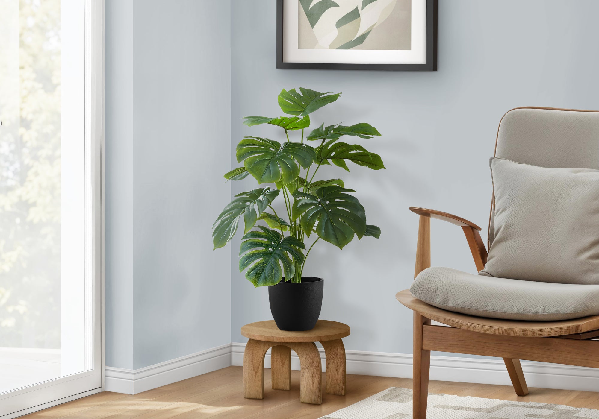 An artificial Medium Monstera plant in a black pot on a small wooden stool next to an armchair.