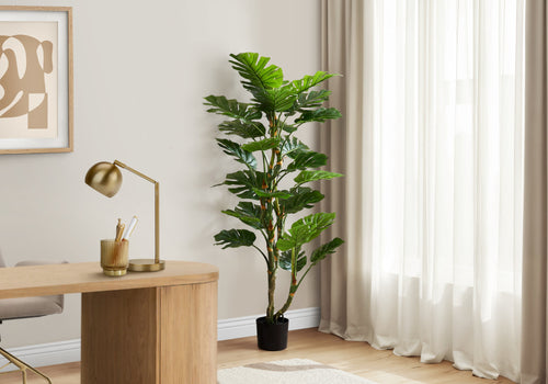 Tall artificial Monstera tree with split leaves and dual trunks standing in a corner next to a wooden desk.